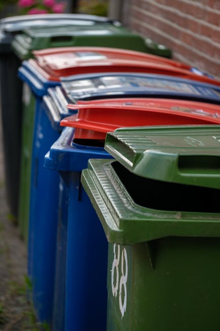 Close-up view of several black plastic rubbish bags filled with waste, tightly knotted at the top and placed against a plain, light-colored wall. The bags have a glossy, reflective surface with visibl