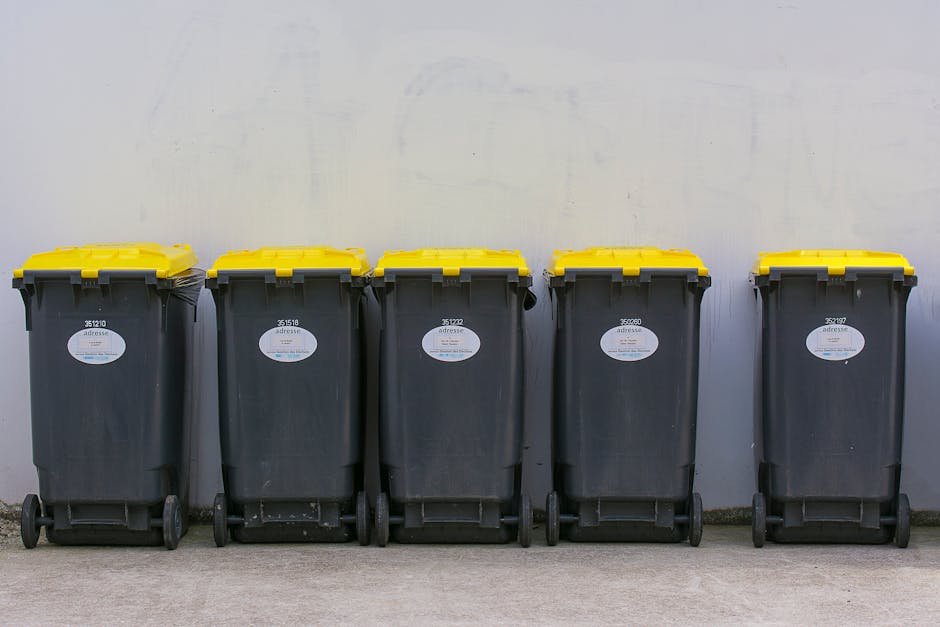 Two large black plastic bags filled with waste are positioned on the pavement beside a black metal fence, which appears to enclose a residential or garden area. The bags are crumpled and slightly miss